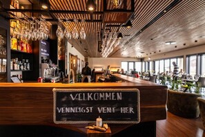 A bar area with a wooden counter, hanging glassware, and a sign that reads 'VELKOMMEN VENNIGST VENT HER'. - Rosenlund Hamn (Sande)