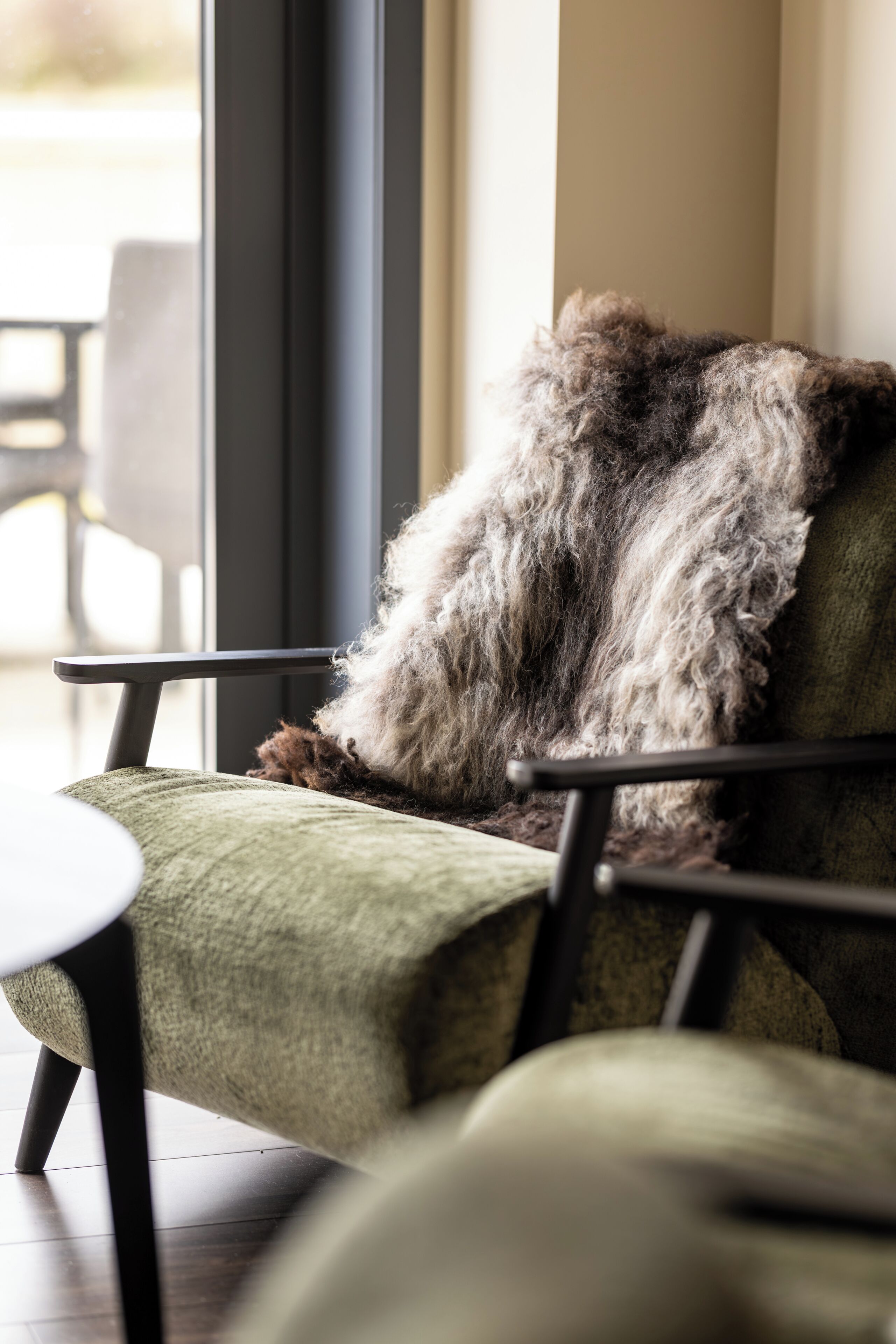 A chair with a fur throw, a round table, and a view of an outdoor area through a window.