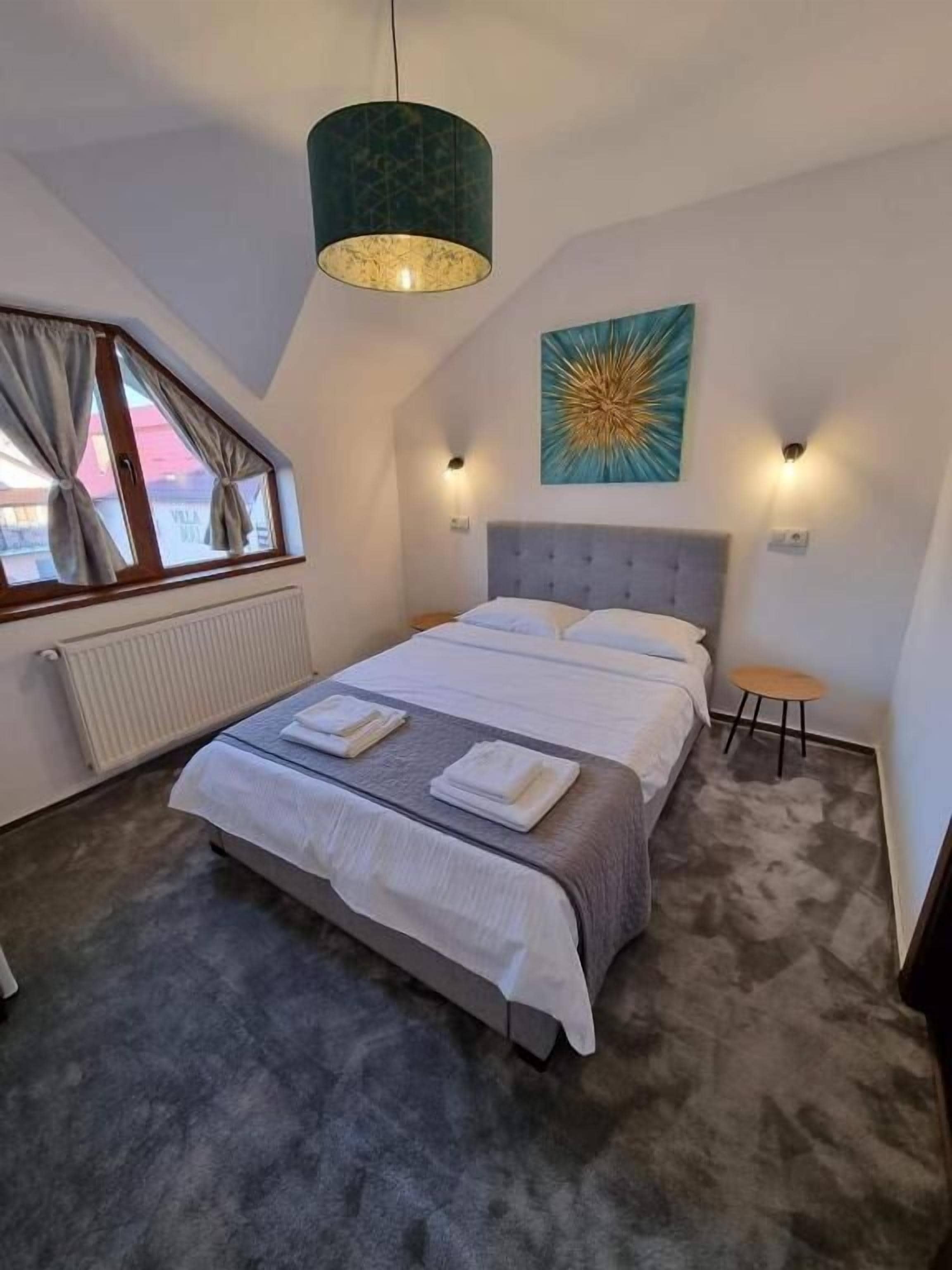 A bedroom with a large bed, a window with curtains, a radiator, a wall-mounted light, and a ceiling light.