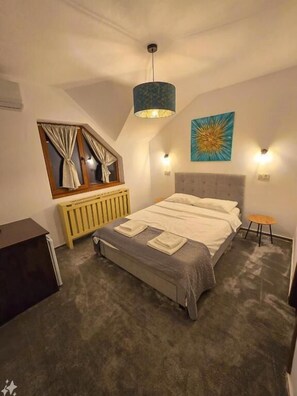 Et soveværelse med seng, et vindue med gardiner, en radiator, en vægmonteret lampe og et billede.