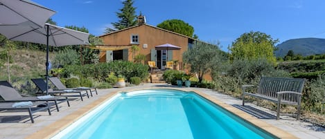 A swimming pool with lounge chairs and umbrellas, a house with a garden, and a mountainous backdrop.