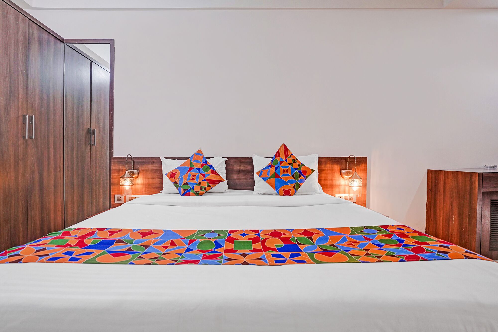 A bedroom with a bed, two pillows, a nightstand, a lamp, and a wardrobe.