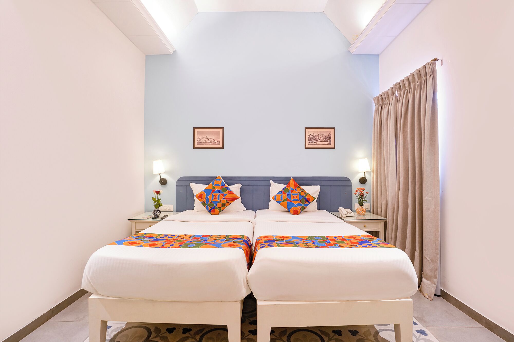 Una habitación de hotel con una cama grande, dos mesitas de noche, una mesita auxiliar, un teléfono, una lámpara, una ventana con cortinas y dos cuadros en la pared.