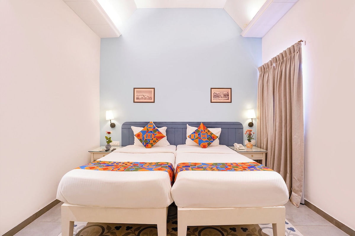 Una habitación de hotel con una cama grande, dos mesitas de noche, una mesita auxiliar, un teléfono, una lámpara, una ventana con cortinas y dos cuadros en la pared.