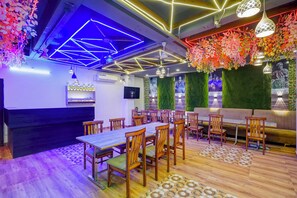 A restaurant with a long wooden table, multiple chairs, and a sofa. There are hanging plants and a wall with a green textured backdrop. - FabHotel SV Grand I - 1 KM From Kashi Vishwanath Temple (Varanasi)