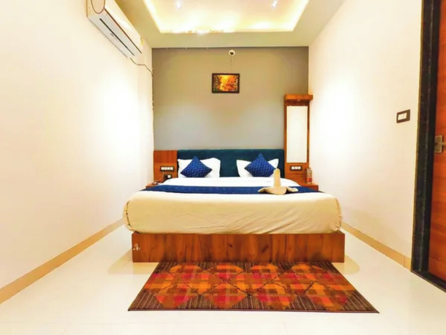 A hotel room with a bed, a headboard, a bedside table, a nightstand, a wall-mounted air conditioning unit, a painting, and a patterned rug.