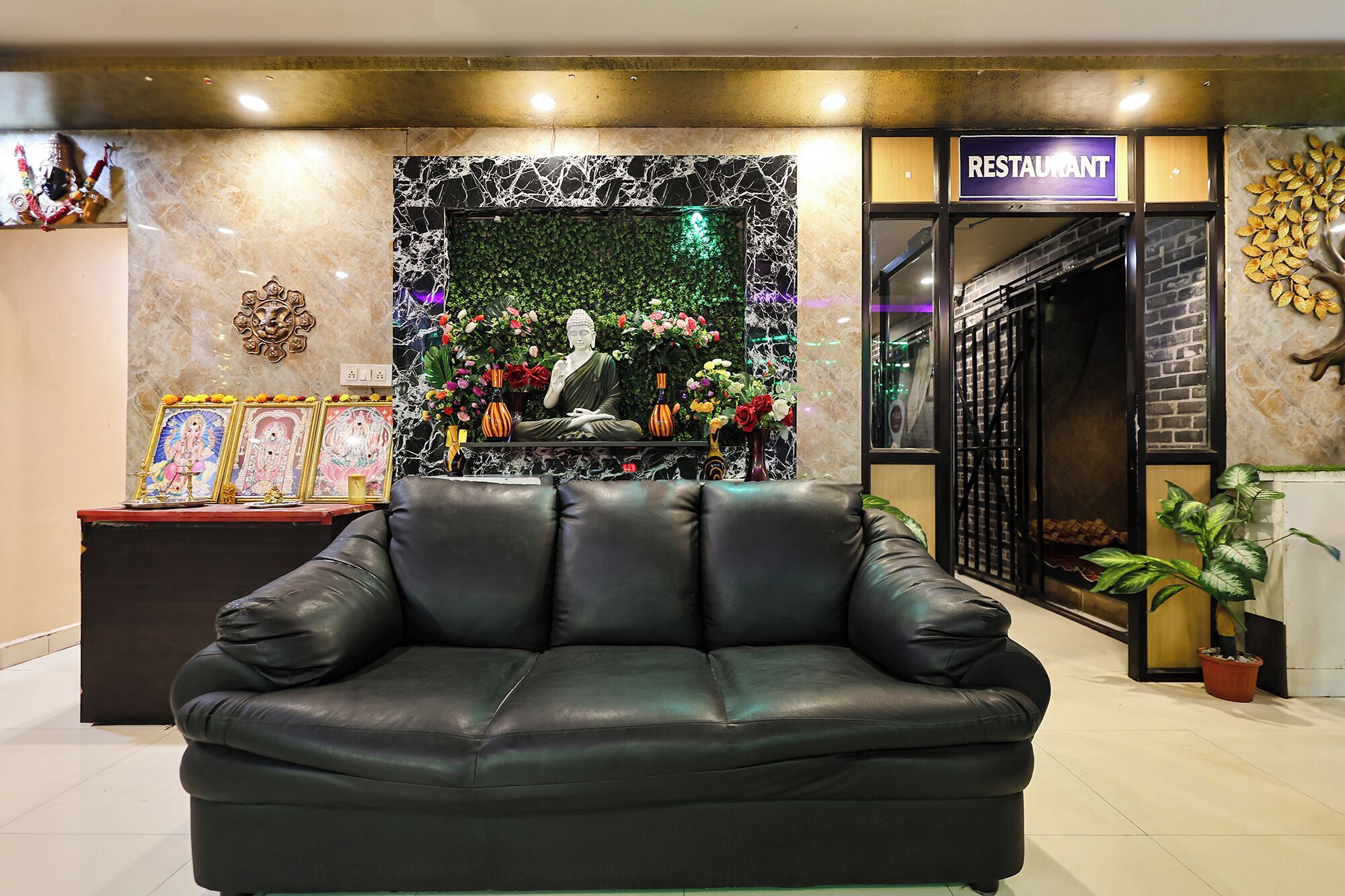 A lobby area with a black leather sofa, a Buddha statue, framed religious pictures, and a restaurant sign.