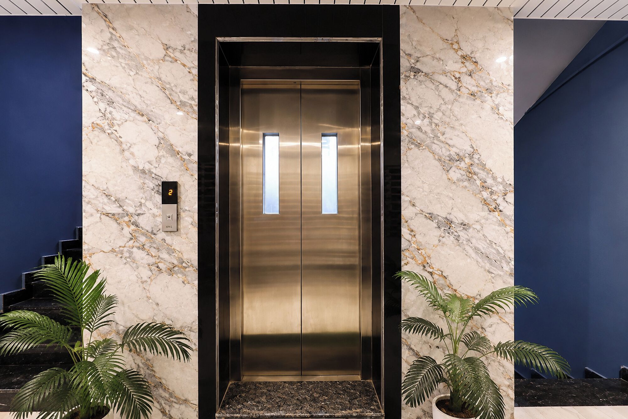 An elevator with a metallic finish, flanked by marble walls and blue accents.