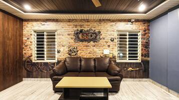 Una habitación con pared de ladrillo, dos ventanas con contraventanas, un sofá de cuero, una mesita y un ventilador de techo.