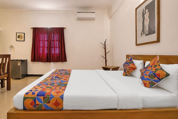 A hotel room with a bed, a nightstand, a chair, a small table, and a wall-mounted air conditioner. - FabHotel Bounty Yatra (Arpora)