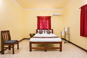 A hotel room with a bed, a chair, a bedside table, a wall-mounted air conditioner, and a window with red curtains. - FabHotel Bounty Yatra (Arpora)