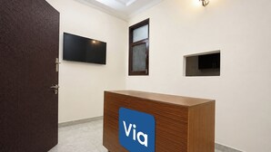 A reception area with a wooden desk, a flat-screen TV, a window, and a wall-mounted light fixture. - Via Platinum Key (New Delhi)