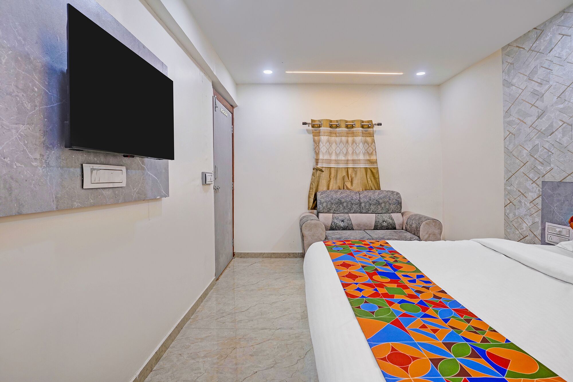 A hotel room with a bed, a television mounted on the wall, a patterned curtain, and a chair.