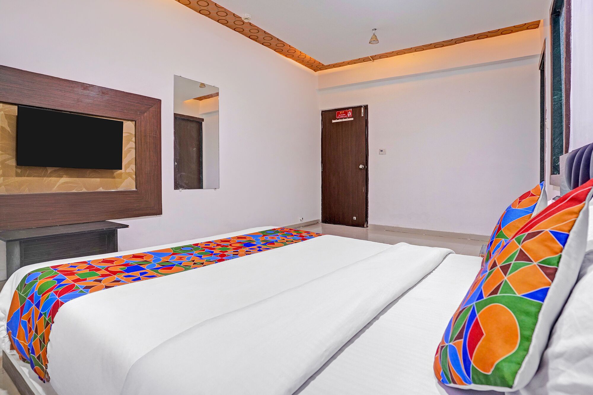 A hotel room with a bed, a television mounted on the wall, a door with a red sign, and a window with curtains.