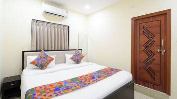 A hotel room with a bed, a nightstand, an air conditioning unit, a wooden door, and a window with curtains.