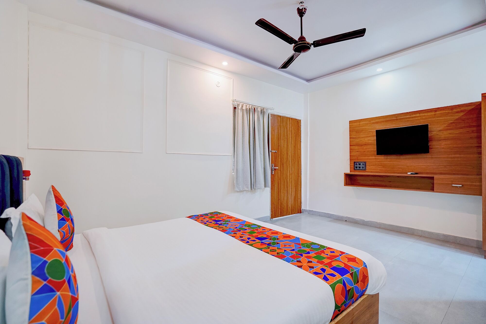 A hotel room with a bed, a ceiling fan, a TV mounted on the wall, and a door with curtains.