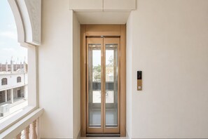 A narrow hallway with a glass elevator door, a wall-mounted elevator control panel, and a window overlooking a building. - Fabhotel Ramanjnaeya (Faizabad)