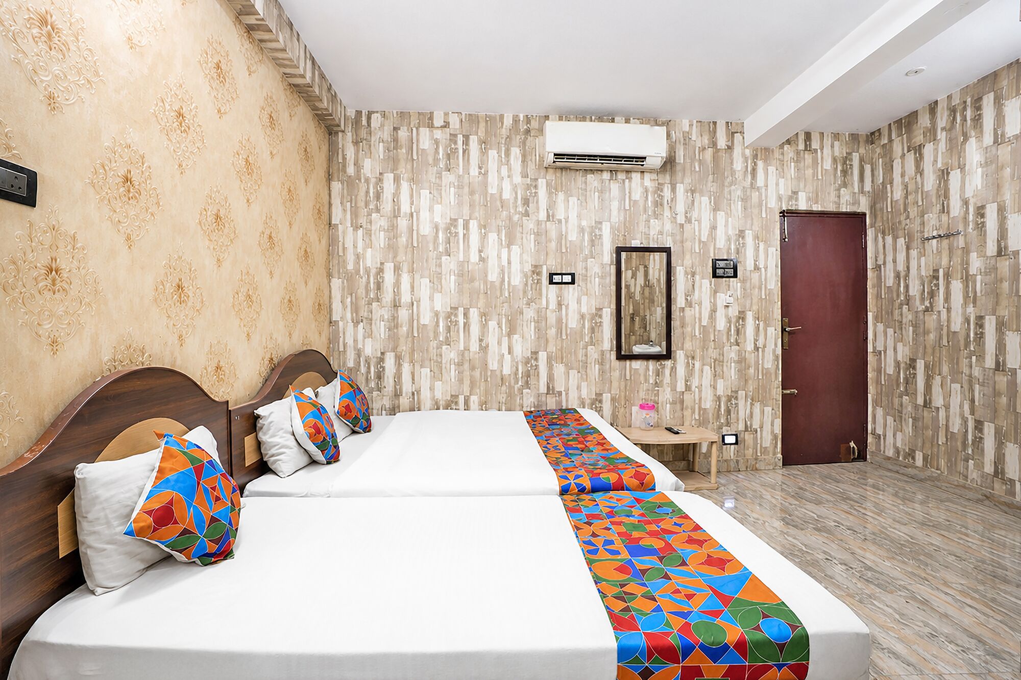 A hotel room with two beds, a wooden headboard, a patterned wall, an air conditioning unit, a door, and a small table with a remote control.