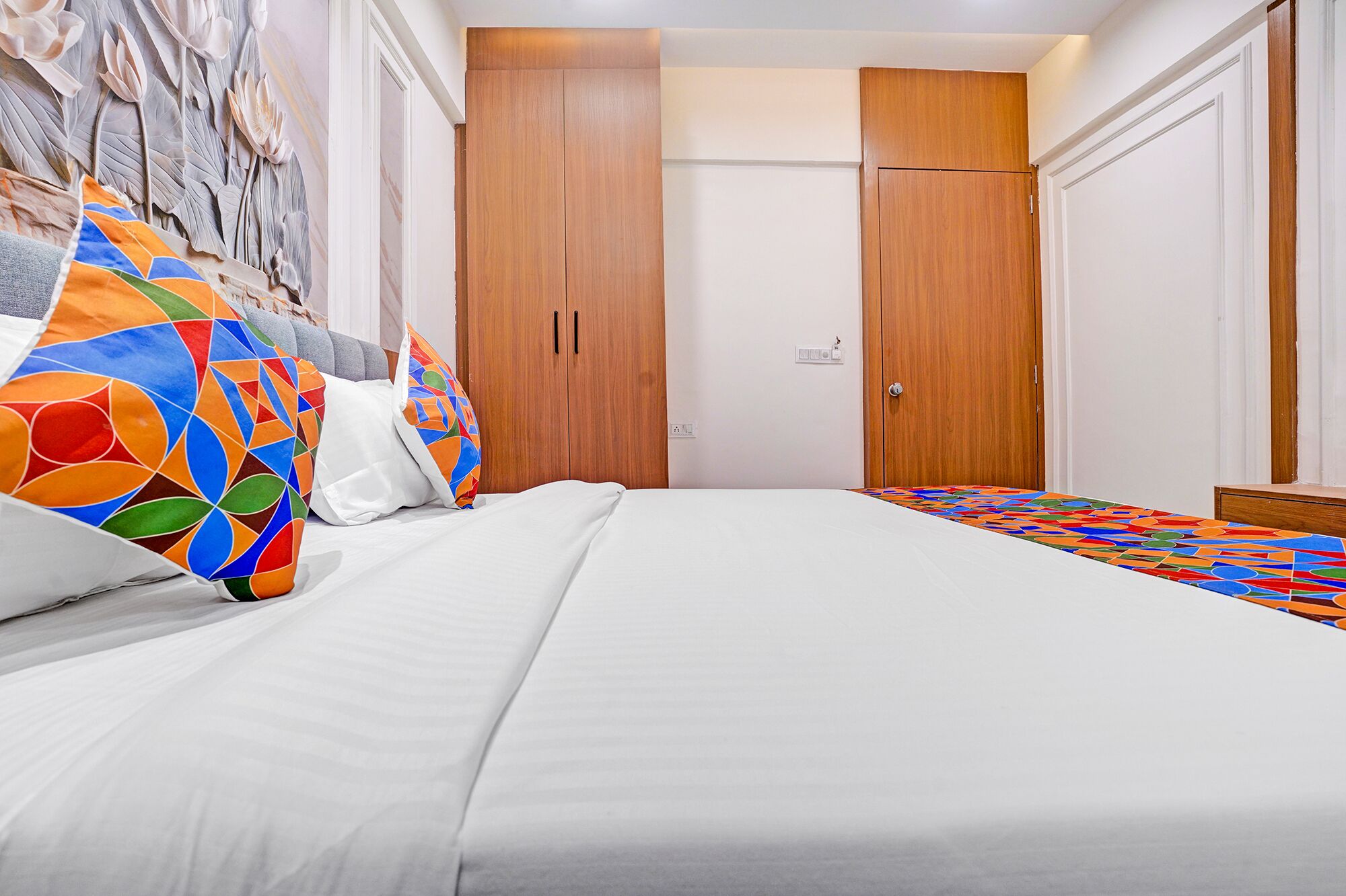 A neatly made bed with white linens, a colorful geometric-patterned pillow, and a wooden wardrobe with a door.