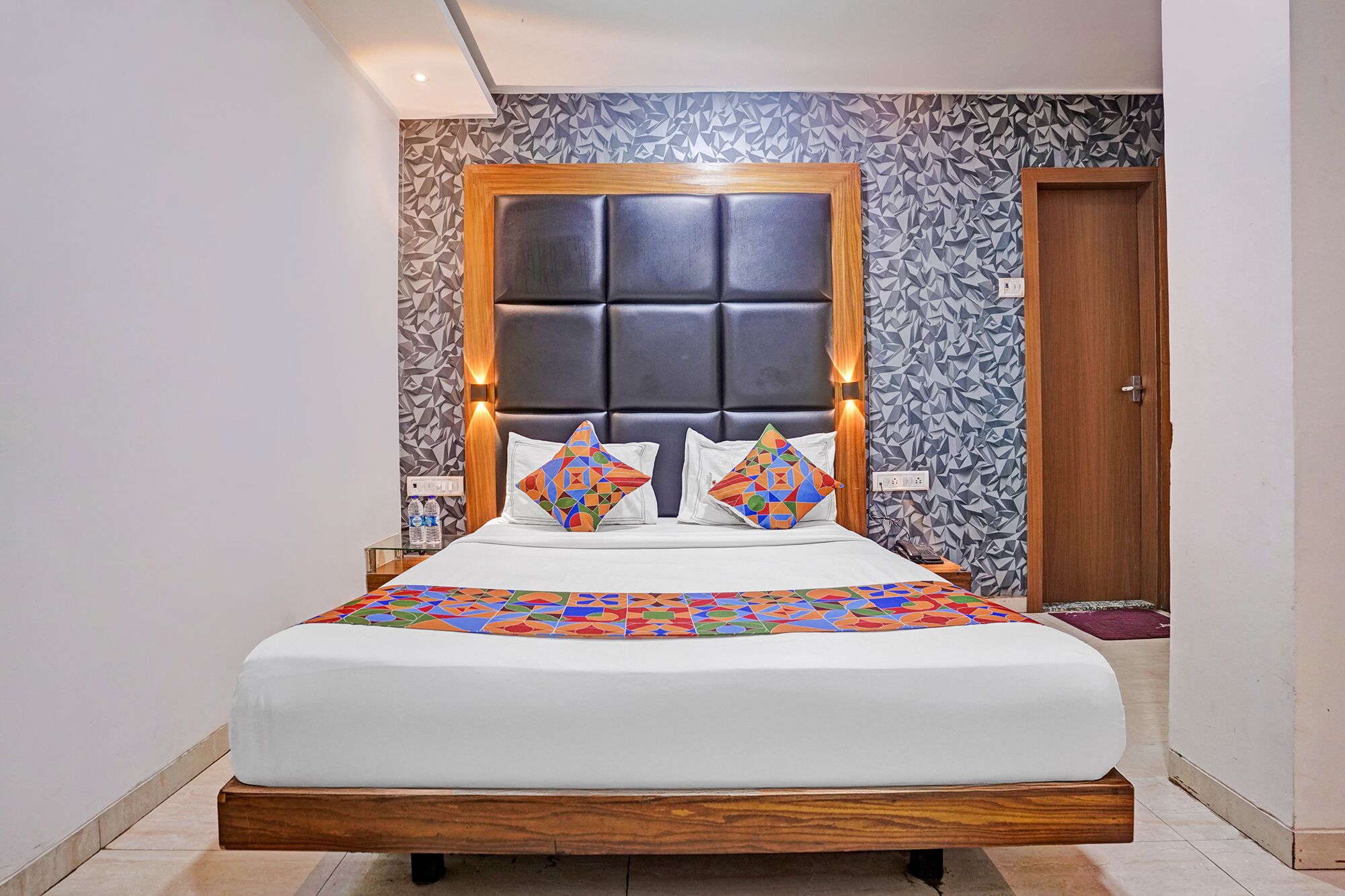 A modern hotel room with a large bed, geometric patterned wallpaper, and a wooden headboard.