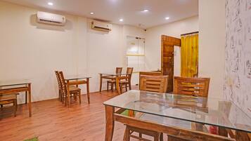 A dining area with a glass-top table and wooden chairs, air conditioning units on the wall, and a yellow curtain.