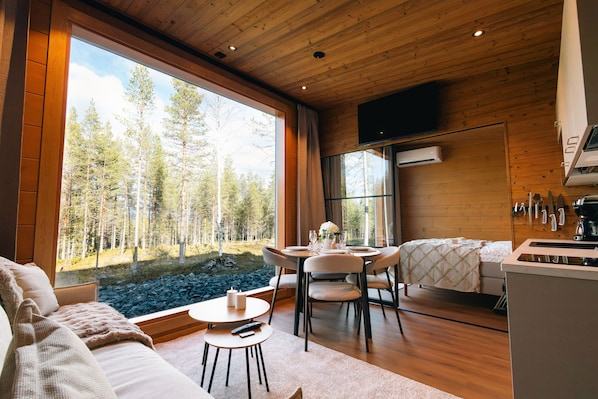 A modern interior with a large window offering a forest view, a dining area, and a kitchen with wooden finishes.