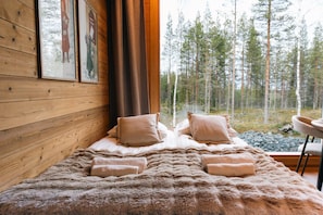 A bed with a fluffy blanket and pillows, a wooden wall with framed artwork, and a window with a forest view.