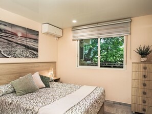 Ein Schlafzimmer mit einem Bett, einem Nachttisch, einem Fenster mit Blick auf Bäume und einer Wand-Klimaanlage.