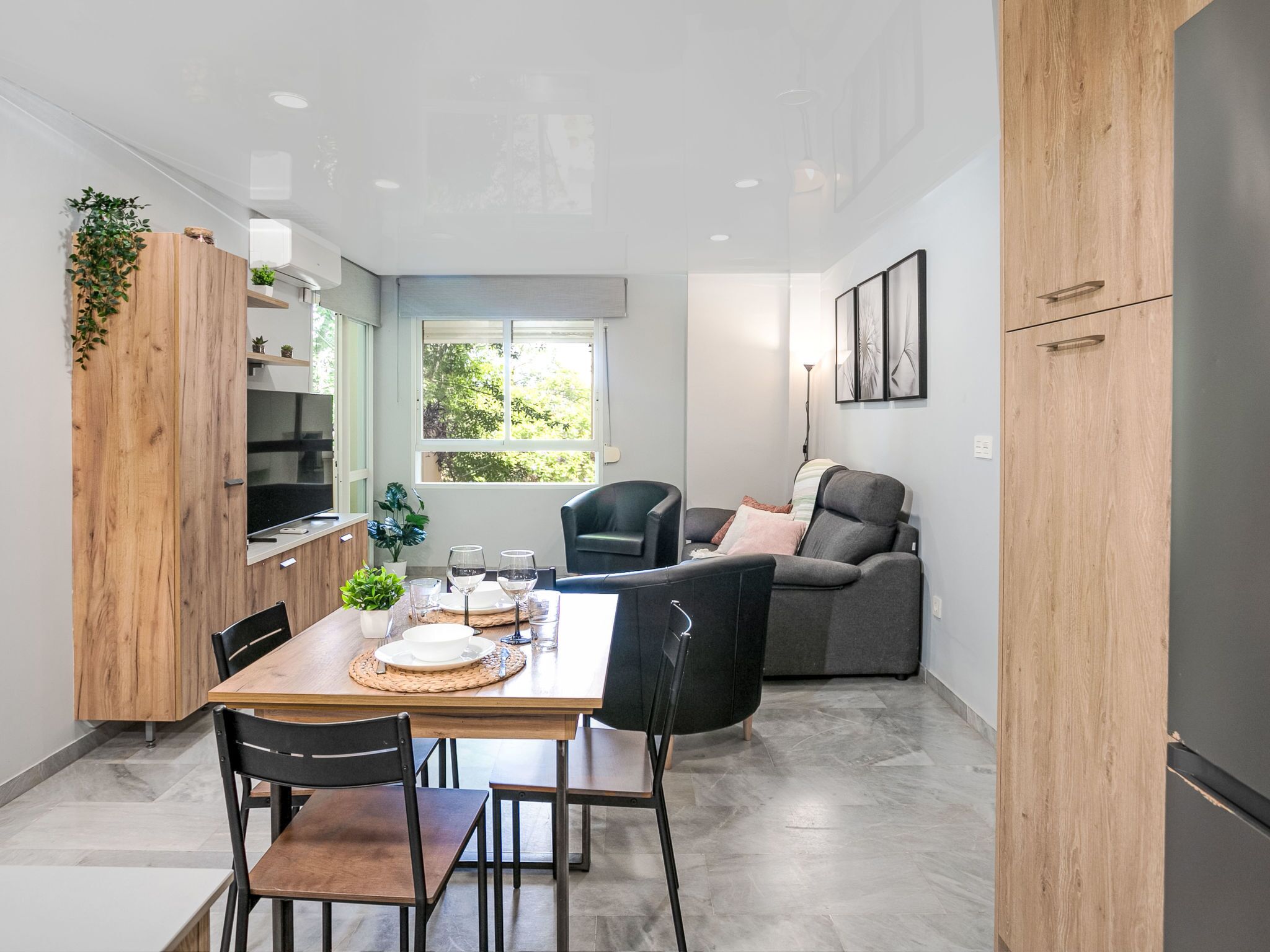 A modern kitchen with a dining area, a sofa, and a window with a view of greenery.