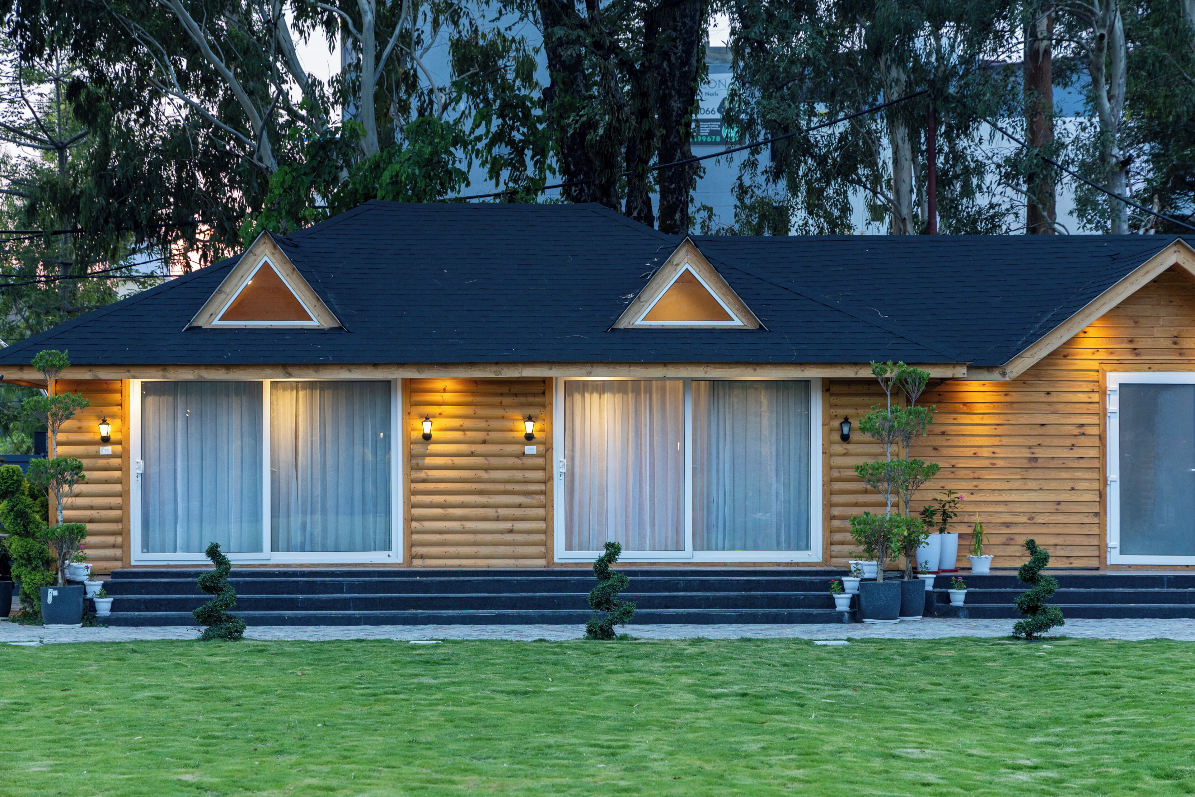 A log cabin with a front porch, two windows with curtains, and a small garden with potted plants.