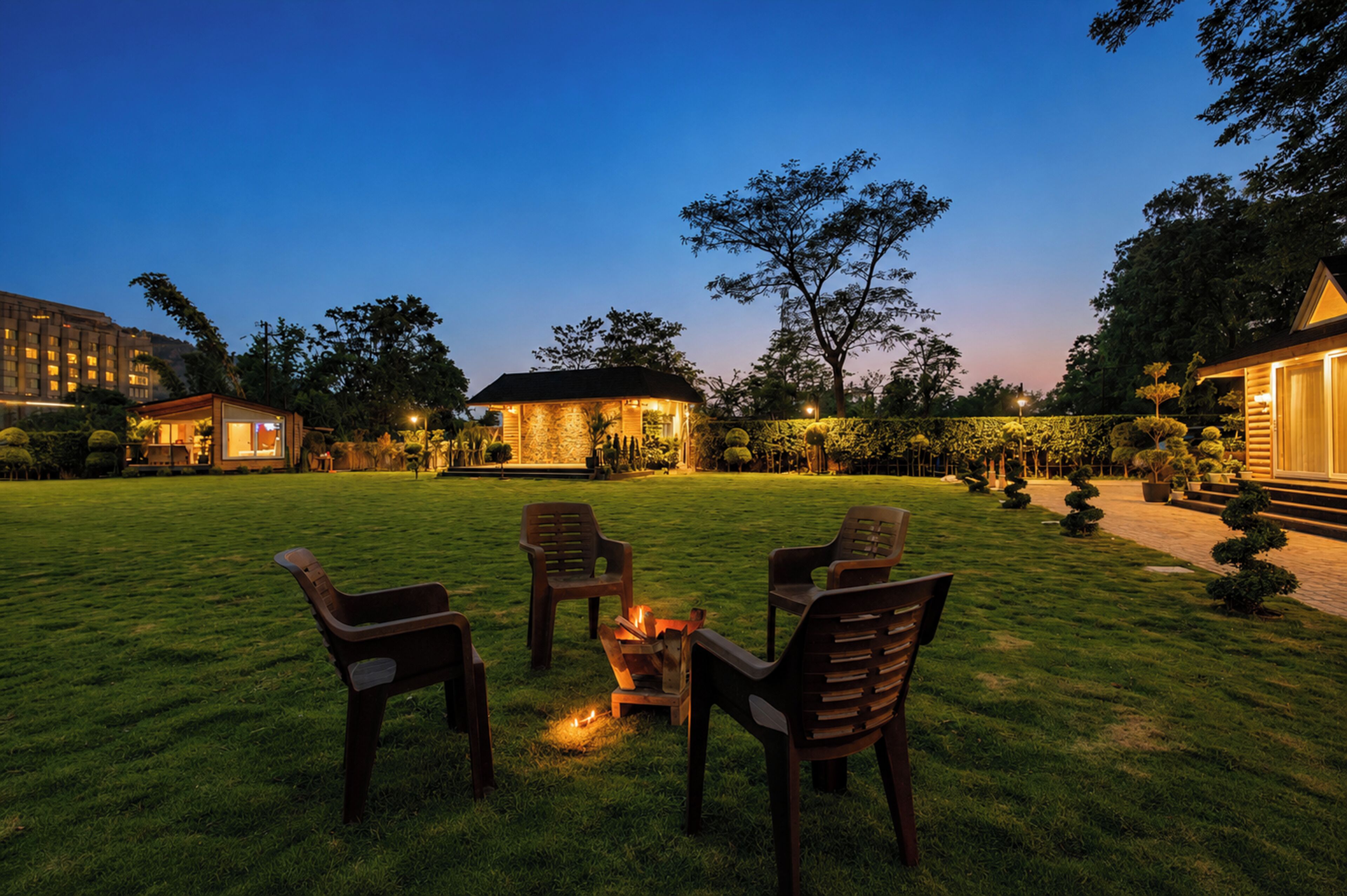 A well-lit outdoor seating area with chairs and a fire pit, surrounded by a manicured lawn and trees.