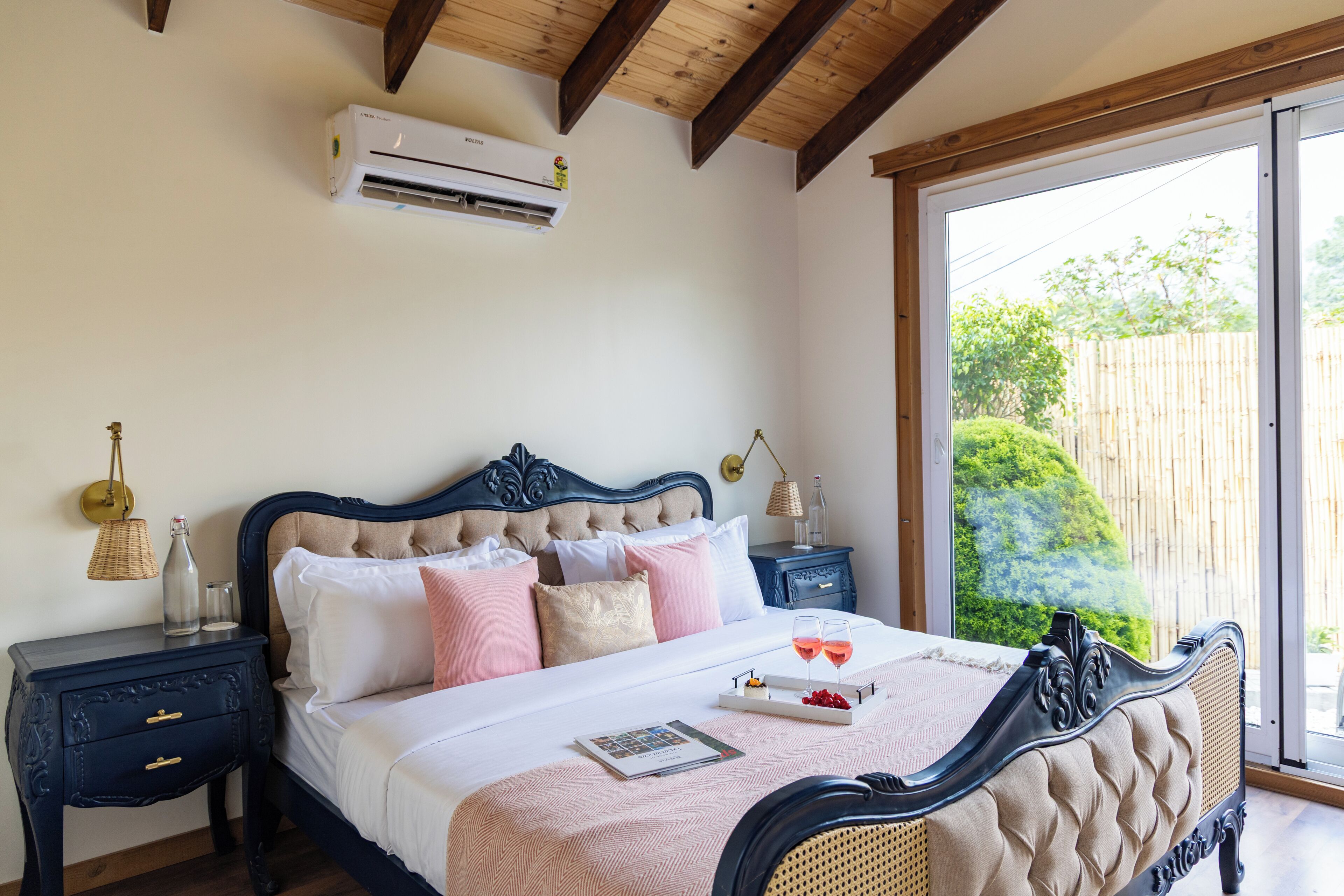 A bedroom with a bed, bedside tables, an air conditioning unit, and a view of greenery outside.