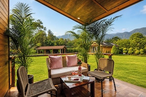 A covered outdoor seating area with a sofa, chairs, and a table, overlooking a well-maintained lawn and distant buildings. - Alaya stays Alora in the hills (Dehradun)