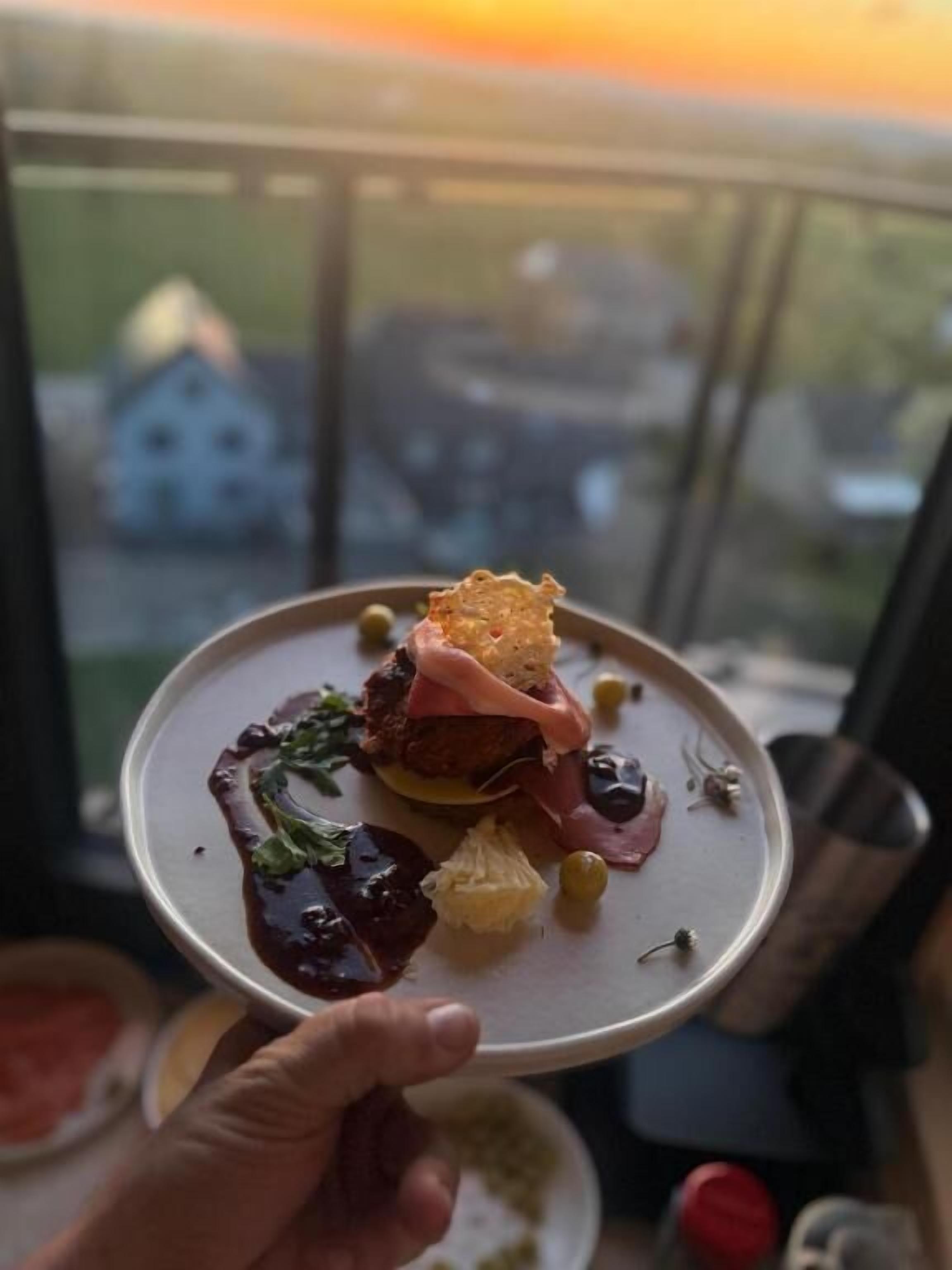 Una persona sosteniendo un plato con comida, una ventana con vista a casas y una barandilla de balcón.