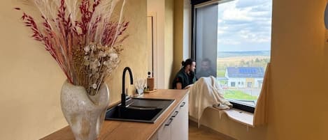 Una cocina moderna con un ventanal grande, un florero con plantas secas y vistas al campo.