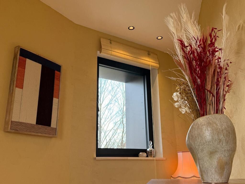 Une pièce avec une fenêtre, un vase contenant des plantes séchées et une œuvre d’art encadrée accrochée au mur.