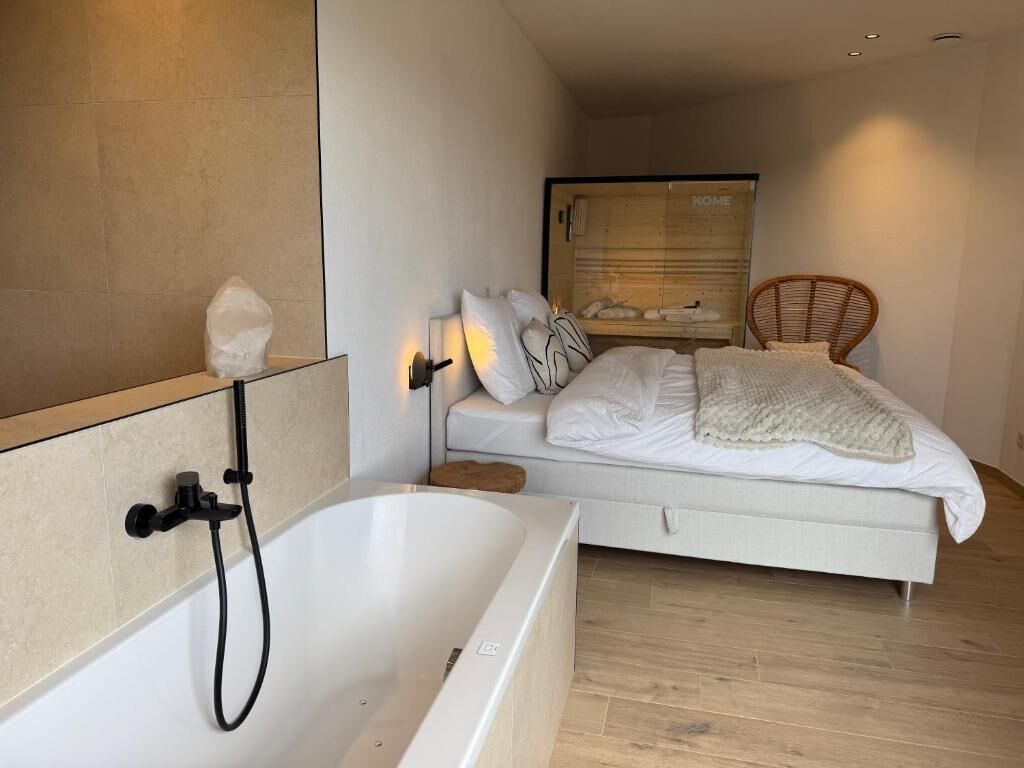 A modern bedroom with a white bed, a wooden headboard, a white bathtub with a showerhead, and a wicker chair.