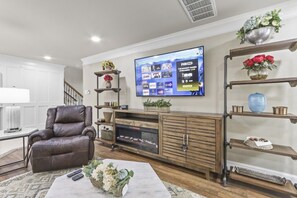 A living room with a leather sofa, a coffee table with decorative items, a flat-screen TV mounted on the wall, and wooden shelves with various decorative objects.