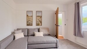 Une chambre avec un canapé-lit gris, deux tableaux accrochés au mur, une porte donnant sur un espace extérieur et une fenêtre avec des rideaux.