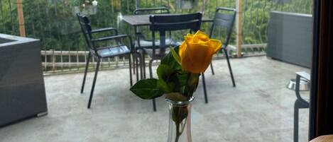 A balcony with a table and chairs, a vase with a yellow flower, and a view of a forested hill.