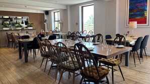 A dining area with wooden tables and chairs, a kitchen in the background, and a large window providing natural light. - Afan Mountain Lodge (Port Talbot)
