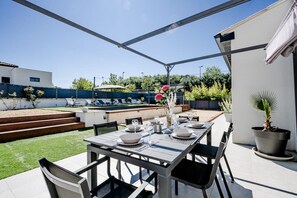 An outdoor patio with a dining table set for four, surrounded by chairs, with a pool and greenery in the background.