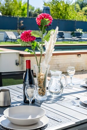 Un espacio de comedor al aire libre con una mesa dispuesta para dos personas, que incluye una botella de vino, copas, una jarra y un florero con flores.