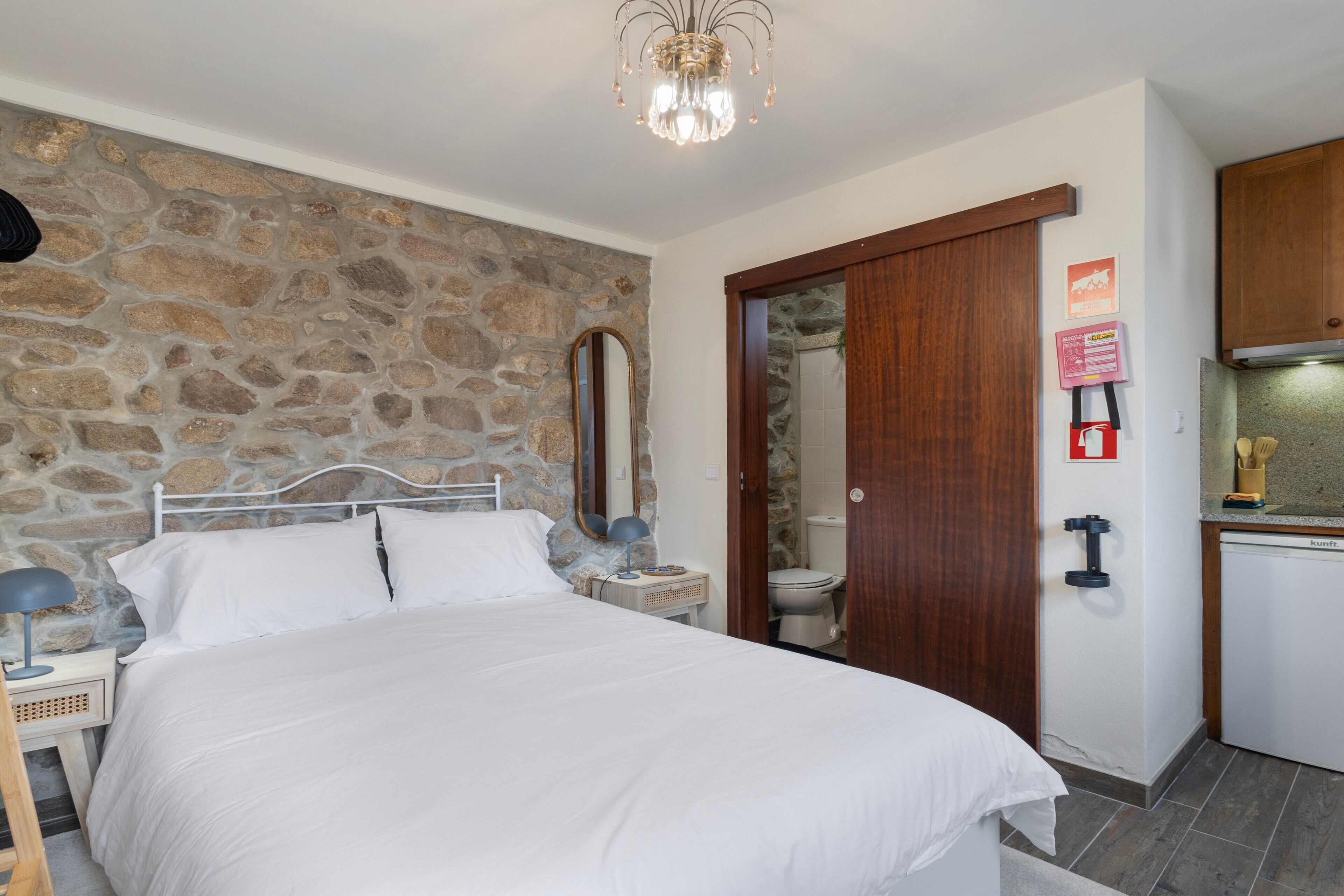 Quarto com parede de pedra, cama com roupa de cama branca, abajur e porta de madeira que dá para um banheiro.
