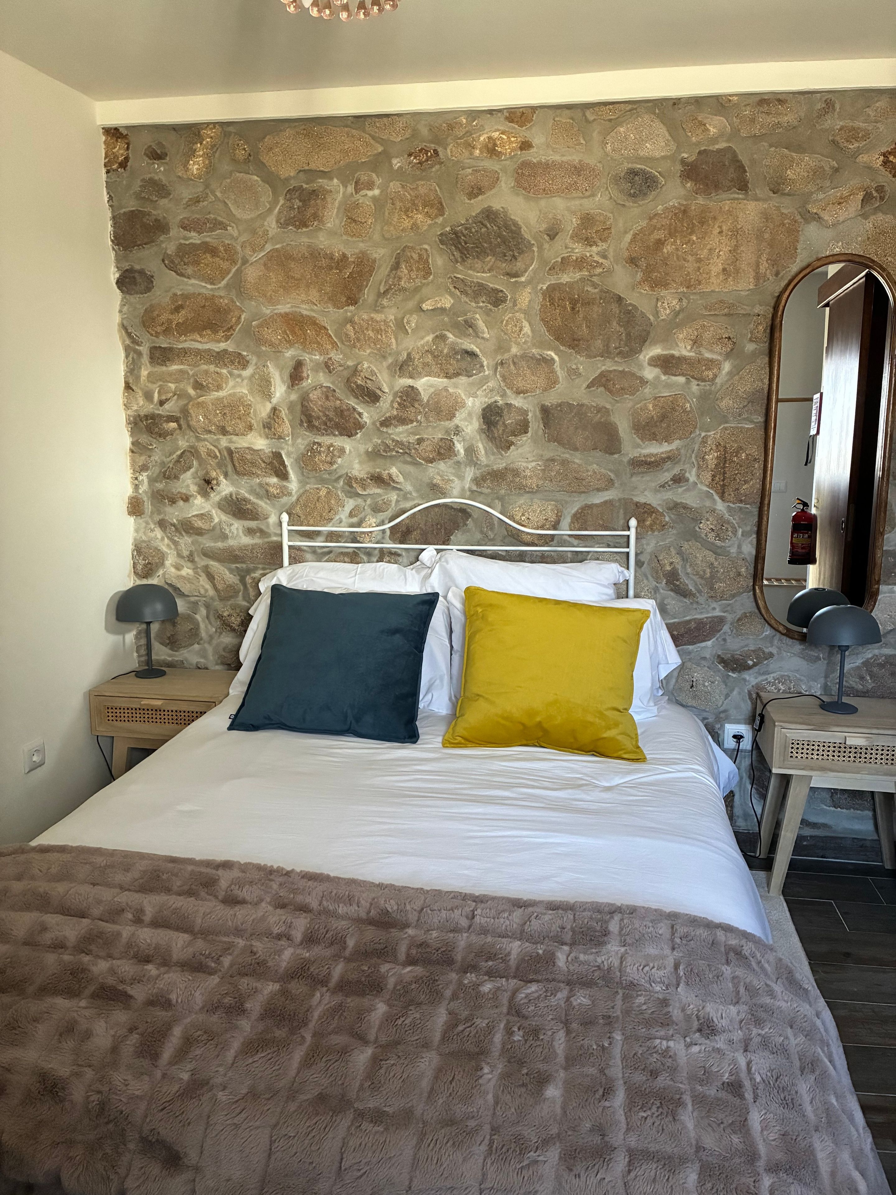 Una cama con ropa de cama blanca y dos almohadas, una pared de piedra al fondo, una mesita de noche de madera con una lámpara y un espejo en la pared.