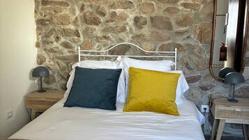 Una cama con ropa de cama blanca y dos almohadas, una pared de piedra al fondo, una mesita de noche de madera con una lámpara y un espejo en la pared.