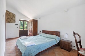 Un dormitori amb un llit, una nitrera de fusta, una cadira i una porta que dona a l'exterior.