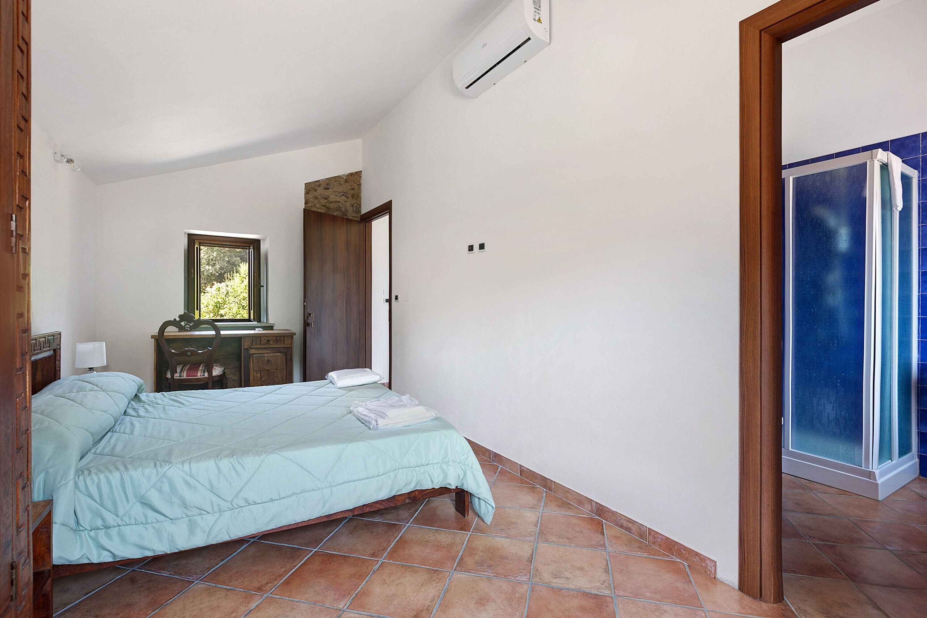 A bedroom with a bed, a desk, a chair, a wardrobe, and a shower area.
