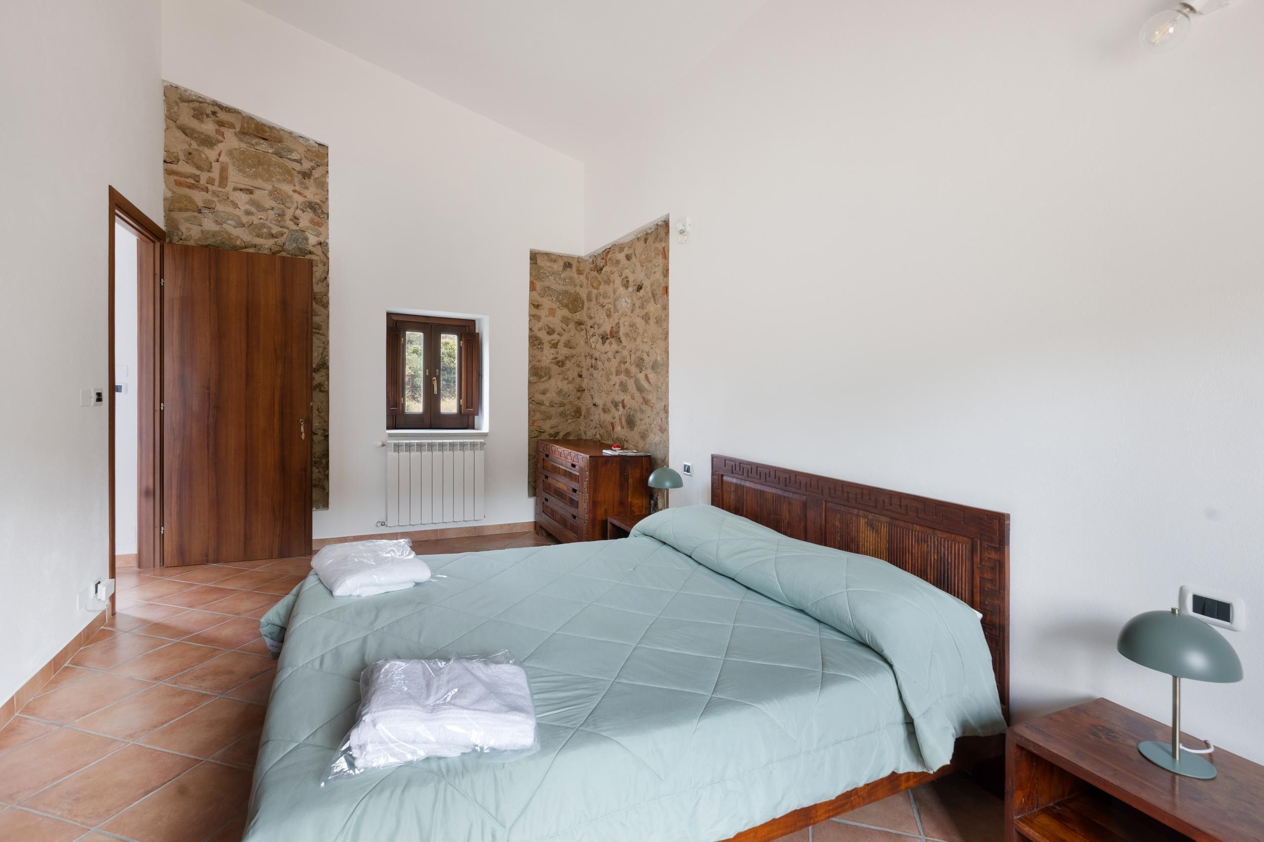 A bedroom with a bed, a wooden wardrobe, a radiator, a window, and a lamp.