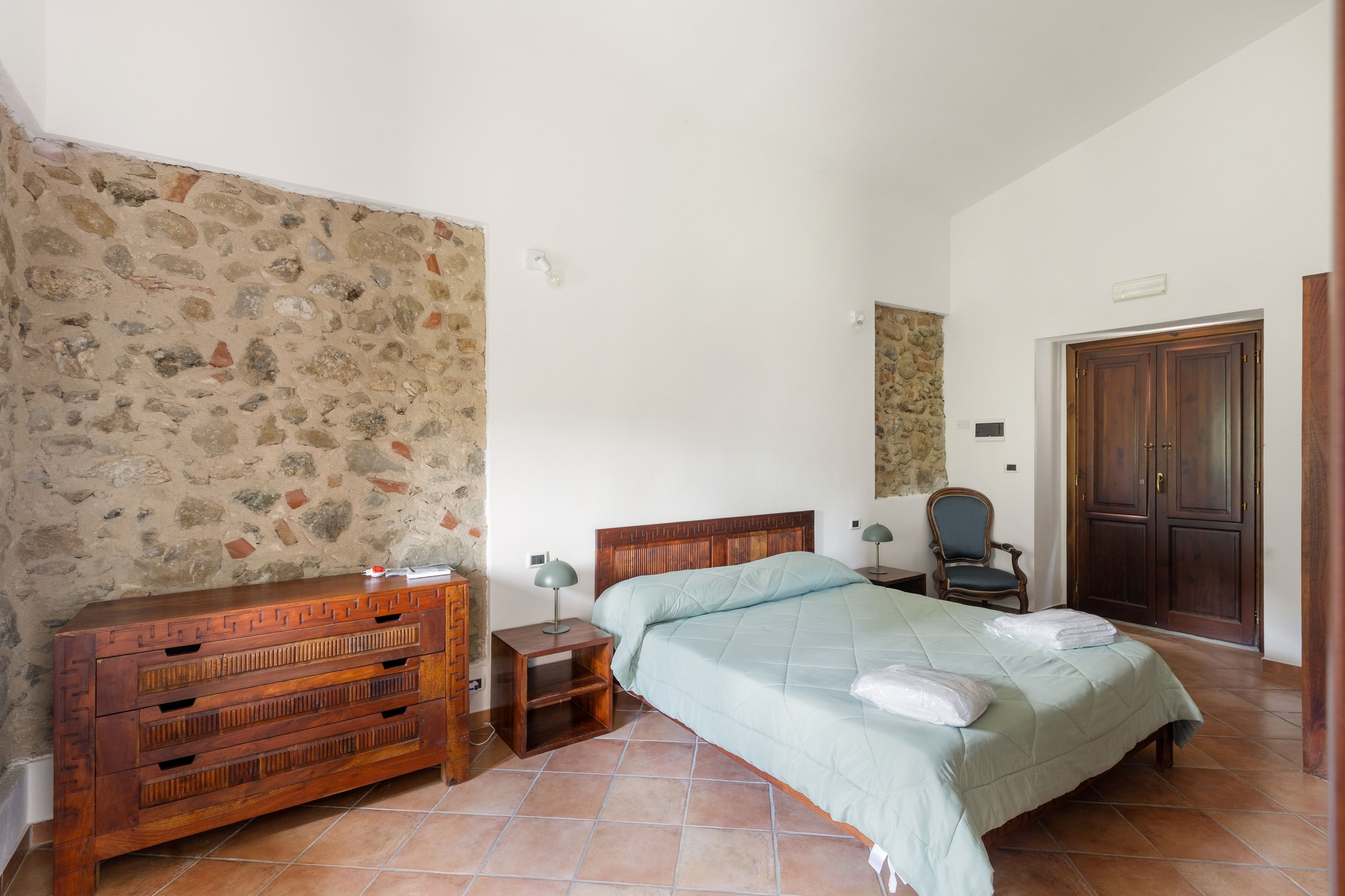 Una camera da letto con parete in pietra, una cassettiera in legno, un letto con copripiumino verde, una sedia e un armadio in legno.